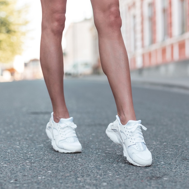 Sportieve slanke vrouwelijke benen in modieuze witte sneakers. Meisje loopt door de stad op asfalt. Close-up van vrouwelijke gebruinde benen in stijlvolle schoenen.