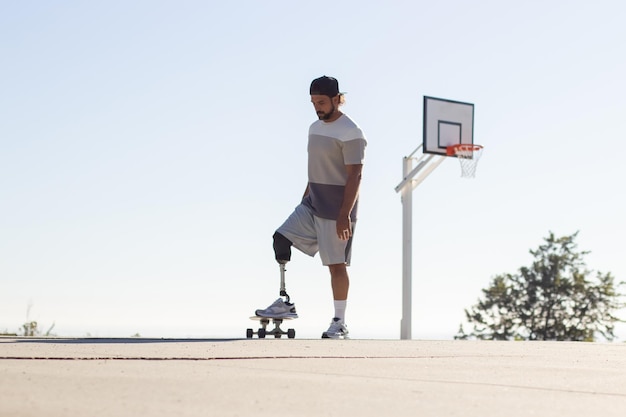 Sportieve stijlvolle man met beenprothese skateboarden in park op zonnige dag. Man die met zijn kunstbeen op skateboard stapt. Basketbalpost op de achtergrond. Geamputeerde sporten, lifestyle concept