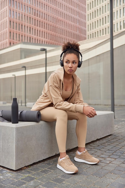 Foto sportieve vermoeide vrouw gekleed in sportkleding luistert naar muziek uit de afspeellijst via een koptelefoon poseert buiten in de buurt van opgerolde fitnessmat en fles geconcentreerd opzij oefeningen in stedelijke omgeving gezonde levensstijl