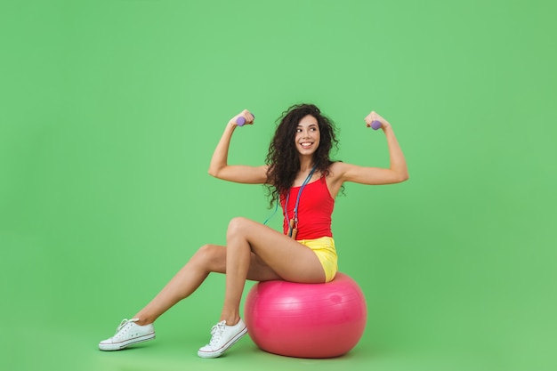 sportieve vrouw die zomerkleren draagt die halters optillen terwijl ze op fitnessbal zit tijdens aerobics tegen groene muur
