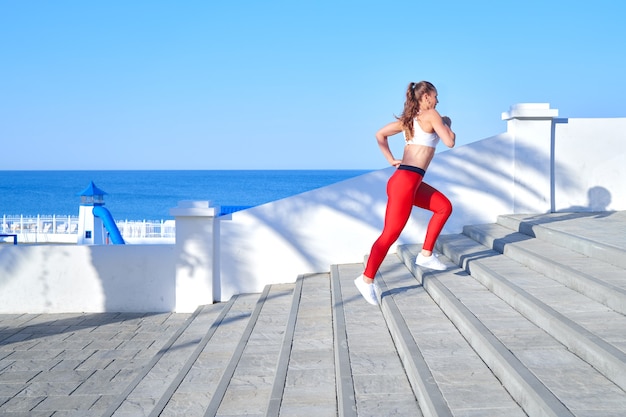 Sportieve vrouw op een zomerochtend joggen op de trap in rode legging op de achtergrond van de zee