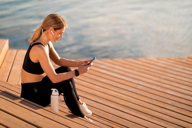 Sportieve vrouw van middelbare leeftijd die smartphone gebruikt terwijl ze ontspant na een training buitenshuis