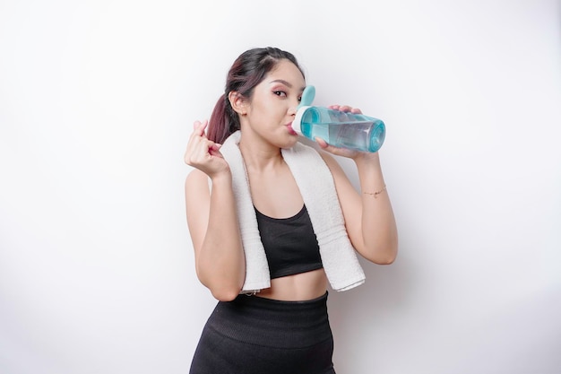 Sportive Asian woman posing with a towel on her shoulder and drinking from a bottle of water smiling and relaxing after workout