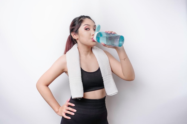 Sportive Asian woman posing with a towel on her shoulder and drinking from a bottle of water smiling and relaxing after workout