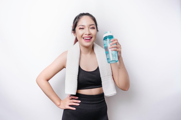 Sportive Asian woman posing with a towel on her shoulder and holding a bottle of water smiling and relaxing after workout