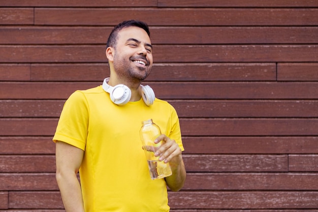 노란색 티셔츠를 입은 스포츠적인 갈색 남자의 초상화 손에 미소 짓는 물병 나무 벽 배경