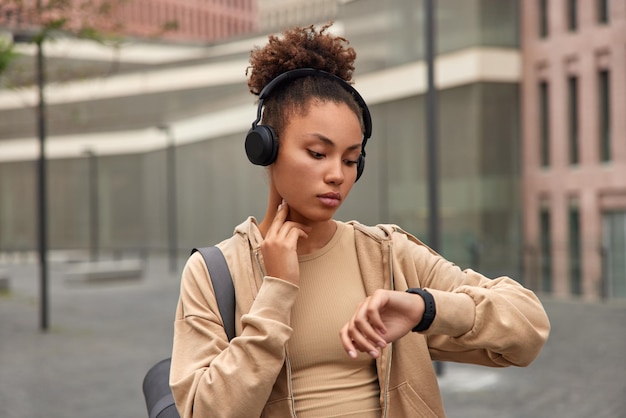 Sportive curly haired young woman checks heart rate pulse looks attenively at smartwatch carries fitness mat and listens music via headphones