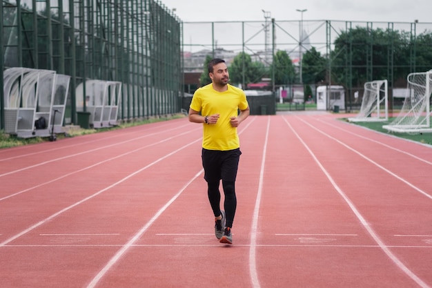 Sportive guy in tracksuit running along red track with white separating lines by soccer field