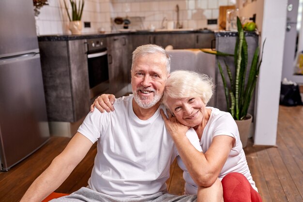 Sportive man and woman 50-60 years old sit on the floor taking a break after workout, sport exercises, look at camera smiling, happy together