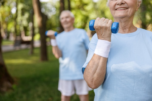 Sportive senior woman with sweatband does exercises with dumbbell training with friend in park