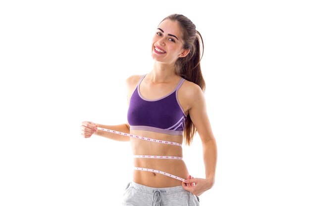 Sportive woman in purple sports top with ponytail using centimeter on white background in studio