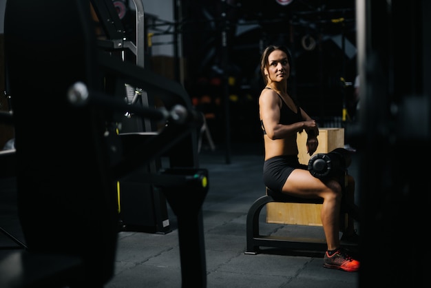 Sportive young woman with perfect athletic body wearing black sportswear sitting at simulator and prepares for training in modern dark gym. Concept of healthy lifestyle.