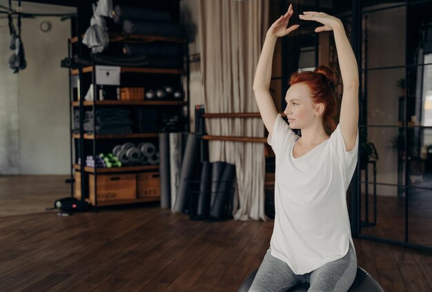 Sportive young woman with red hair sits on big pilates ball with hands lifted over head