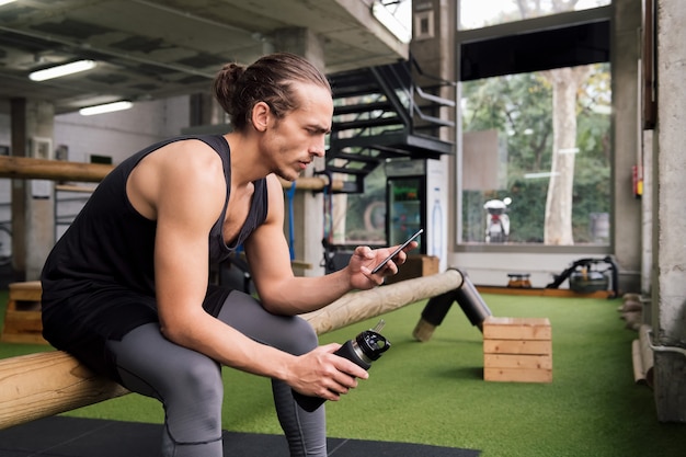 Sportman die een fles vasthoudt, raadpleegt de telefoon in de sportschool