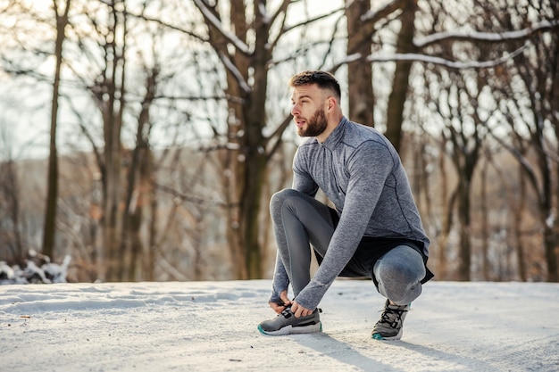 Sportman gehurkt en veter strikken in de natuur op besneeuwde winterdag. Sportkleding, winterfitness, koud weer