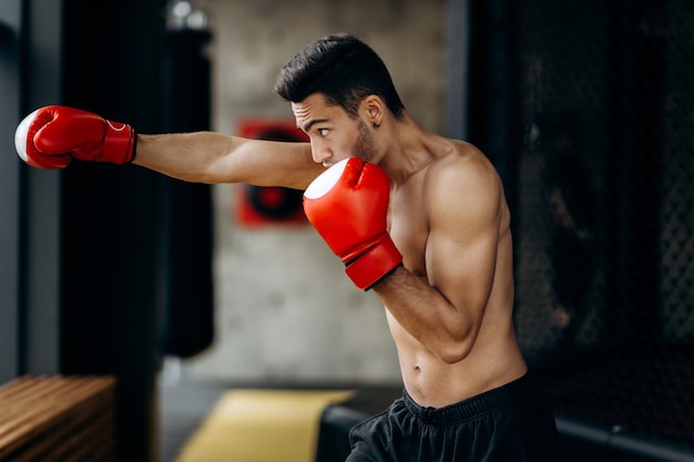 Sportman met een naakte torso en de rode bokshandschoenen aan zijn handen staat hits met de rechterhand in de boksschool.