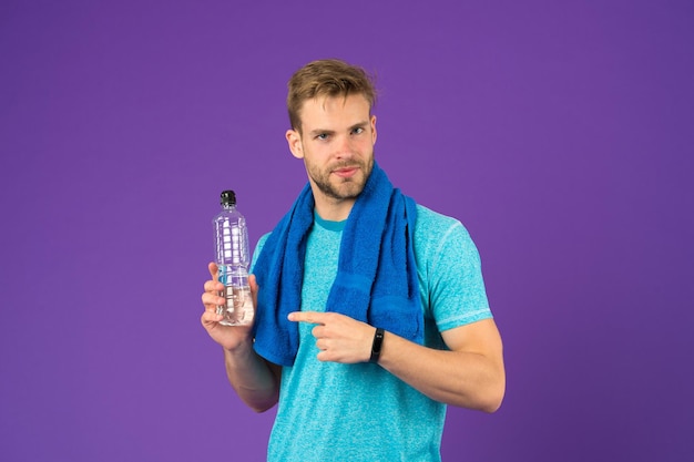 Sportman wijsvinger op plastic fles op violette achtergrond Man houdt waterfles atleet in blauwe t-shirt en korte broek met handdoek na de training Dorst en uitdroging Sportactiviteit en energie