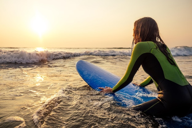 Спортивная красивая женщина в водолазном костюме, лежащая на доске для серфинга в ожидании большой волны. Девушка серфинга в гидрокостюме, серфинг в океане на закате. Мокрые волосы, счастье и свобода, пляжный отдых