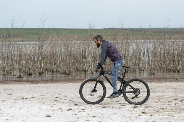 Спортивный брутальный бородатый парень на современном горном велосипеде Велосипедист в соляном безлюдном месте у озера