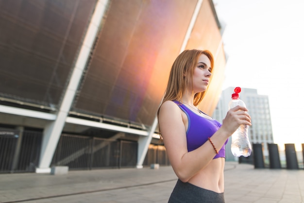 Спортивная девушка в спортивной одежде, стоя перед стадионом с бутылкой воды в руках и смотрит в сторону.