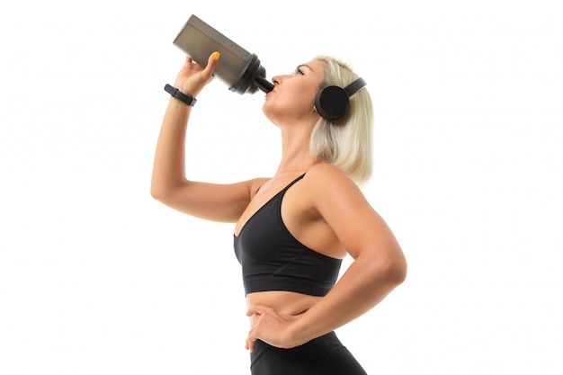 A sports girl with blonde hair and bright manicure in headphones, black sports topics and leggings drinks from a sports water bottle.