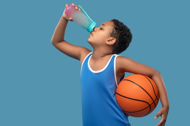 Sports, thirst. dark skinned boy sideways in blue shirt with\
ball drinking water from sports bottle