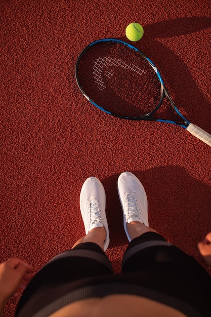 Sports woman athlete getting on outdoor red hardcourt in\
summer. sporting event concept. professional player preparing for\
tournament. top view of feet in white sneakers and a tennis\
racket