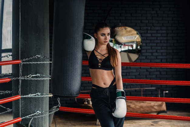 Sports woman in black sportswear and white boxing gloves have a rest after hard training in modern loft gym. Sporty lifestyle concept.