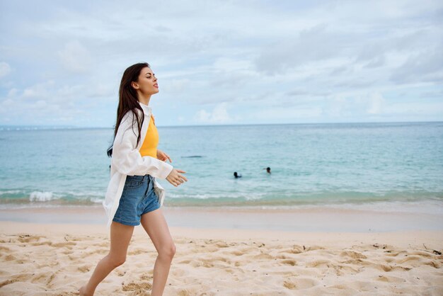 スポーツ女性は、黄色のタンクトップとデニムのショートパンツの砂の上に夏服を着てビーチに沿って走り、白いシャツに髪を飛ばし、オーシャンビューのビーチでの休暇と旅行の自由を楽しみます