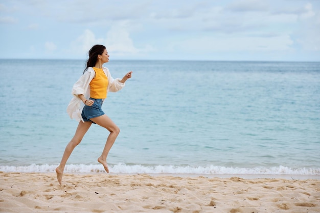 Спортивная женщина бежит по пляжу в летней одежде по песку в желтой майке и джинсовых шортах, белой рубашке с развевающимися волосами, видом на океан, пляжным отдыхом и путешествиями
