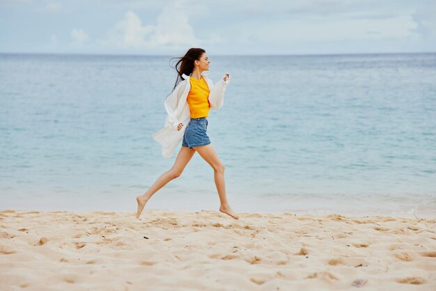 Sports woman runs along the beach in summer clothes on the sand in a yellow Tshirt and denim shorts white shirt flying hair ocean view