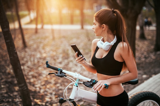 ヘッドフォンでスポーツの女性は電話で立っているし、公園で日没で自転車の近くの距離に外を見て