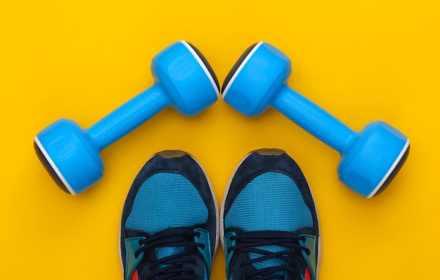 Sportschoenen (sneakers) en blauwe halters op een gele achtergrond. Gezonde levensstijl, fitnesstraining. Bovenaanzicht