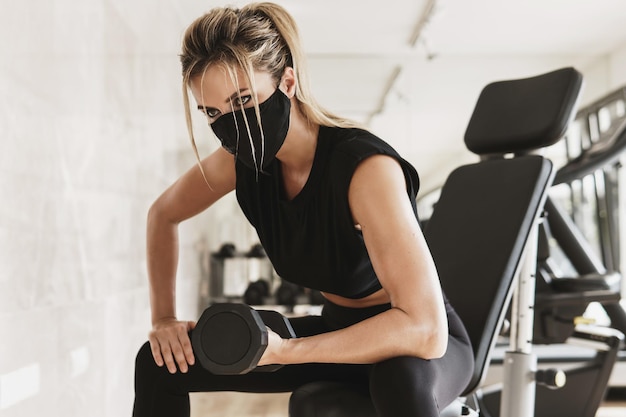 Sportschool nieuw normaal. Jonge atletische vrouw die een preventief gezichtsmasker draagt tijdens haar fitnesstraining met dumbbells.