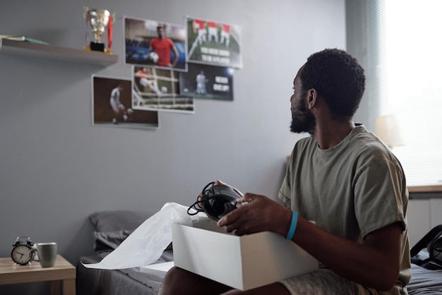 Sportivo di etnia africana che guarda i poster dei giocatori di football sul muro mentre è seduto sul letto di casa