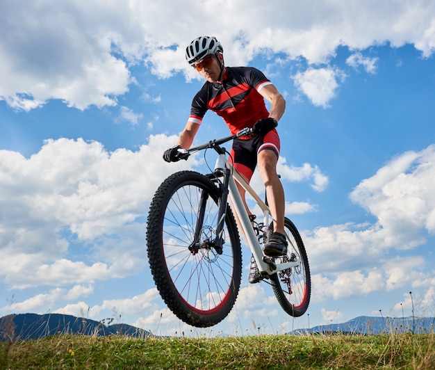 白い雲と遠くの丘の背景と明るい青空に彼の自転車で空を飛んでいるスポーツマンバイカー