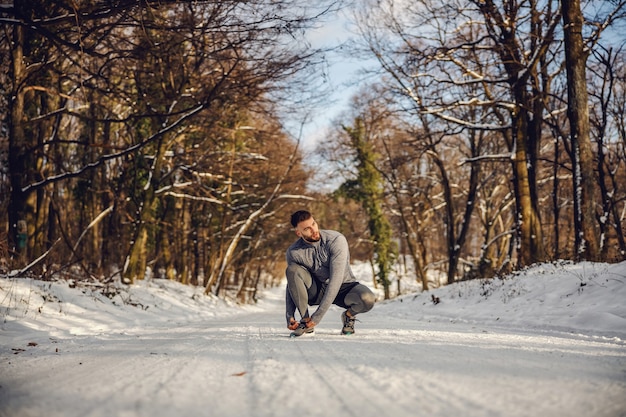 Sportsman crouching and tying shoelace in nature at sunny snowy winter day. Sportswear, winter fitness, cold weather