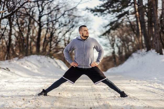 Sportsman doing splits and stretching exercises while standing in nature at snowy winter day