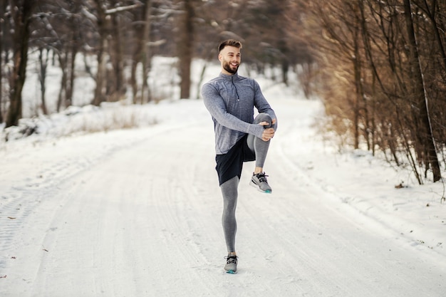雪の降る冬の日にストレッチ体操をし、自然の中で走る準備をしているスポーツマン。冬のフィットネス、スポーツ、寒さ