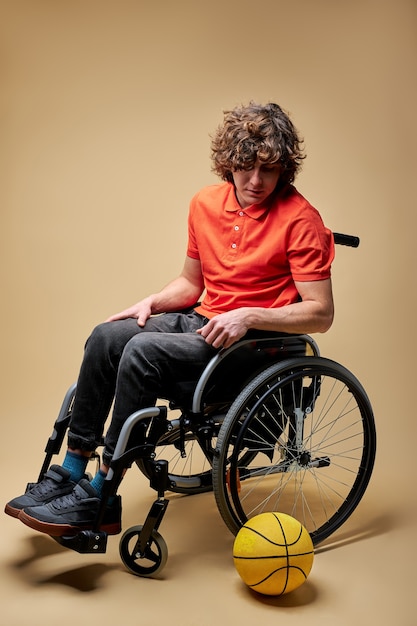 Sportsman had to give up sports due to disability, he sits on wheelchair, looking at ball with sad expression on face.