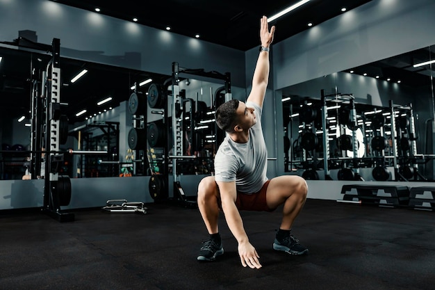 A sportsman squatting and doing stretching exercises for arms in a gym