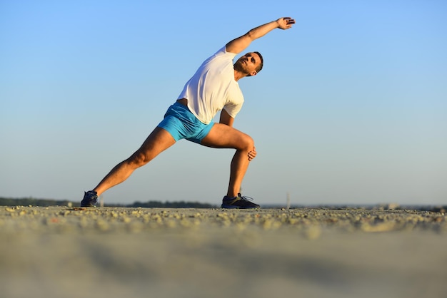 Lo sportivo indossa una maglietta bianca e pantaloncini blu che allungano i muscoli. stile di vita sano e concetto di sport. l'uomo con la figura sportiva fa yoga. il trainer fa esercizi di riscaldamento sullo sfondo del cielo blu.