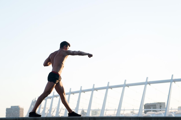 Sportsman with muscular body on white sky. Man doing boxing exercises on roof. Athlete training outdoors. Fitness and sport. Healthy lifestyle concept.