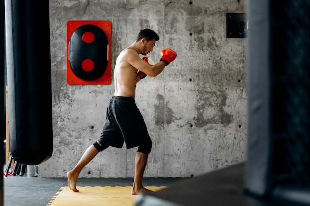 Sportsman with a naked torso and in the red boxing gloves hits equipment for boxing on a concrete wall in the gym .