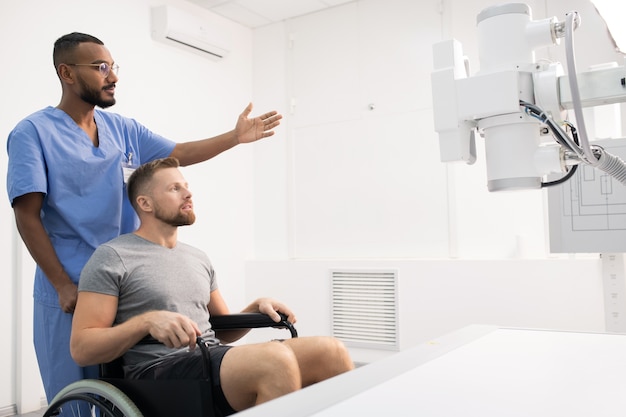 Sportsman with sick leg sitting on wheelchair in front of medical equipment while his doctor pointing at it