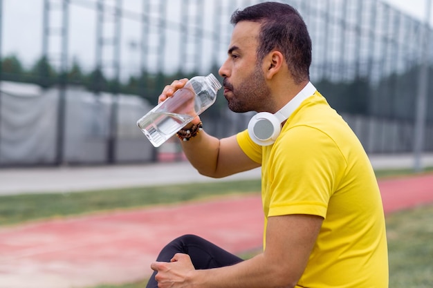 Photo sportsman with wireless headphones drinking fresh water from transparent bottle to refresh