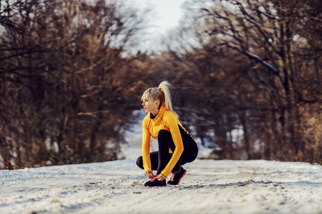 冬の自然の中で雪道にしゃがみ、靴ひもを結ぶスポーツ選手。スポーツウェア、冬のフィットネス、健康的な生活