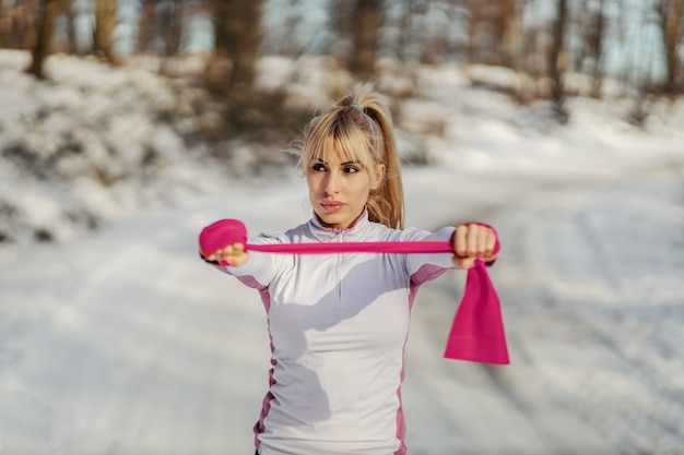 Sportswoman doing fitness exercises with power rubber while standing in nature at snowy winter day. Healthy life, winter fitness, outdoor fitness