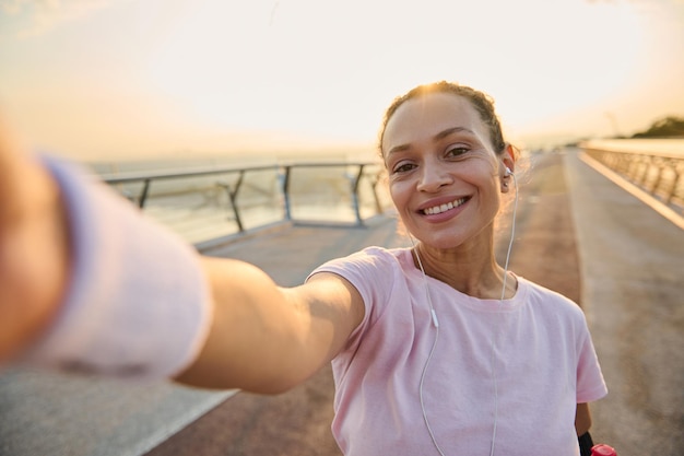 Sportswoman, female athlete in pink t-shirt and wristbands\
smiles holding a mobile phone in her outstretched hands while\
making self-portrait during morning jog and workout on the bridge\
at sunrise