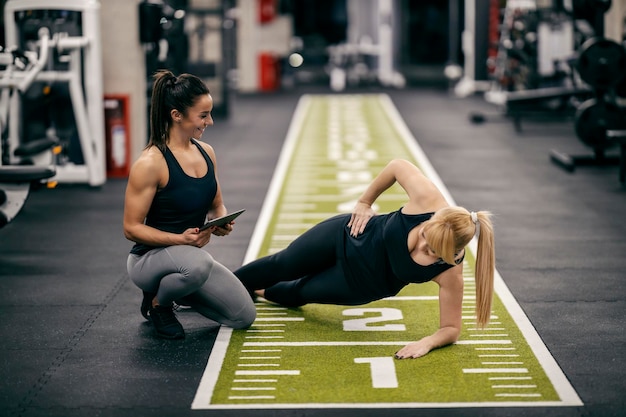 a sportswoman is doing side planks while her female personal trainer is watching her
